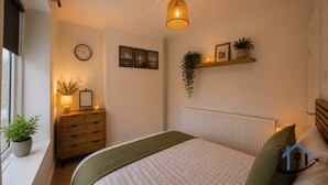 A bedroom with a bed, bedside table, lamp, clock, framed pictures, and a hanging plant. - Barret Flat, sleeps 4, central location, Penarth (Penarth)