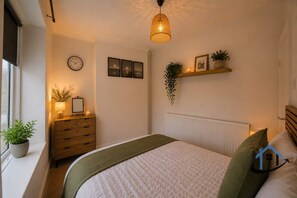 Ein Schlafzimmer mit Bett, Nachttisch, Lampe, Uhr, gerahmten Bildern und einer hängenden Pflanze.