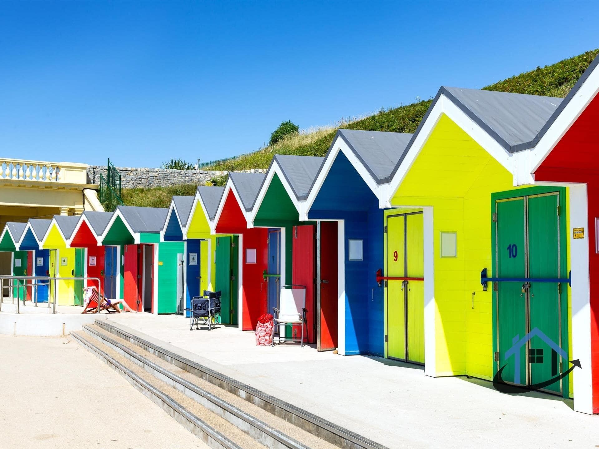 A row of brightly colored beach huts with doors numbered 9 and 10, a chair, and a suitcase on the steps.