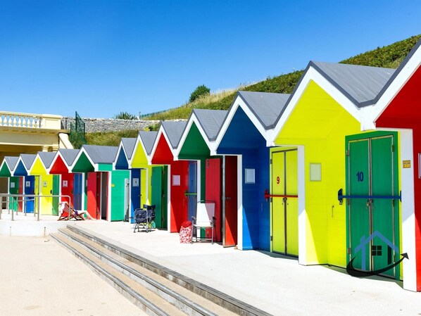 A row of brightly colored beach huts with doors numbered 9 and 10, a chair, and a suitcase on the steps.