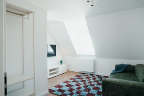 Une pièce moderne et bien éclairée, avec un plafond en pente, un téléviseur à écran plat fixé au mur, un canapé vert et un tapis à motifs.