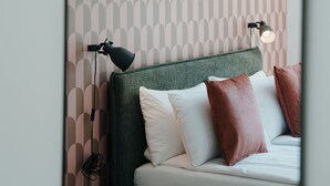 Un dormitorio con papel tapiz estampado, cabecero tapizado en verde, ropa de cama blanca y dos lámparas de noche.