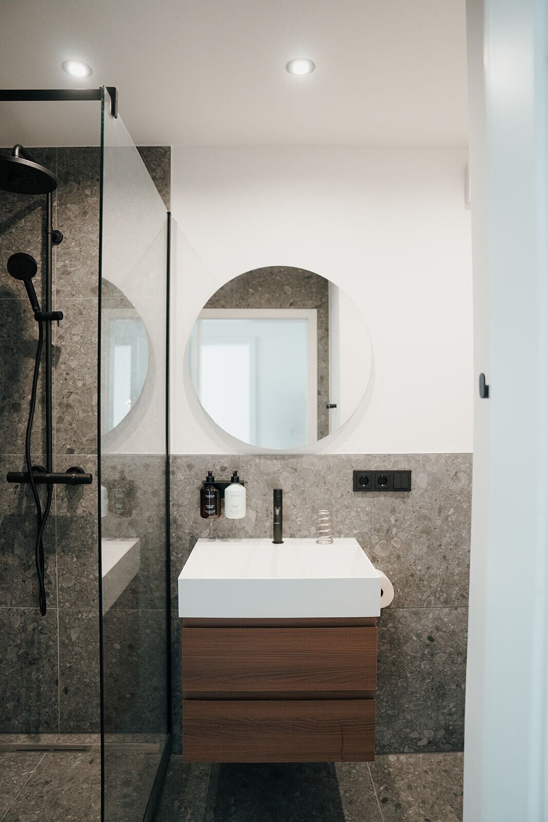 Un baño moderno con cabina de ducha de vidrio, un lavabo blanco con mueble de madera y un espejo redondo sobre el lavabo.