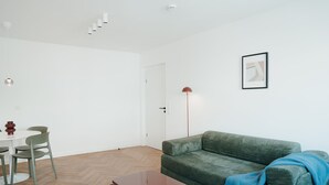 A minimalist living room with a green sofa, a coffee table, a dining table with chairs, and a floor lamp.