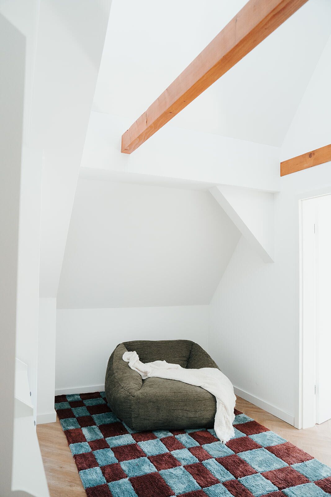 A room with a sloped ceiling, a wooden beam, a white door, and a checkered rug.