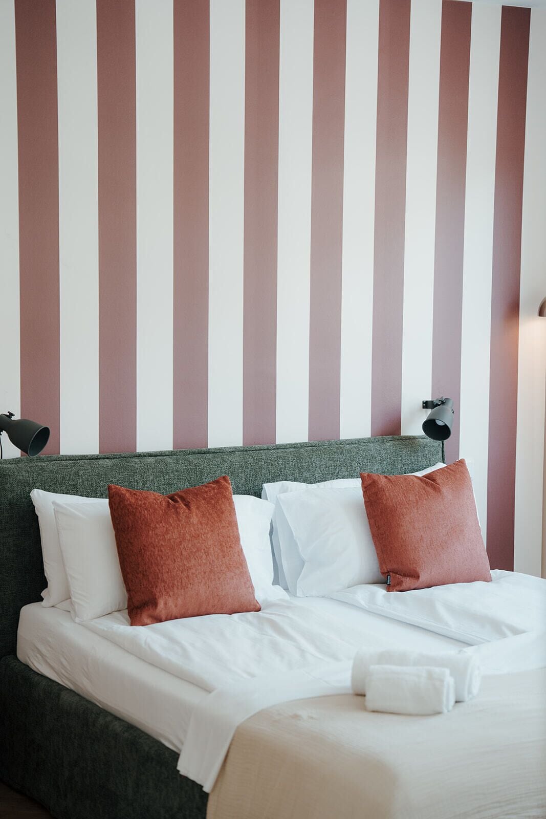 A neatly made bed with white linens and two brown pillows, a green headboard, and a striped wall in the background.