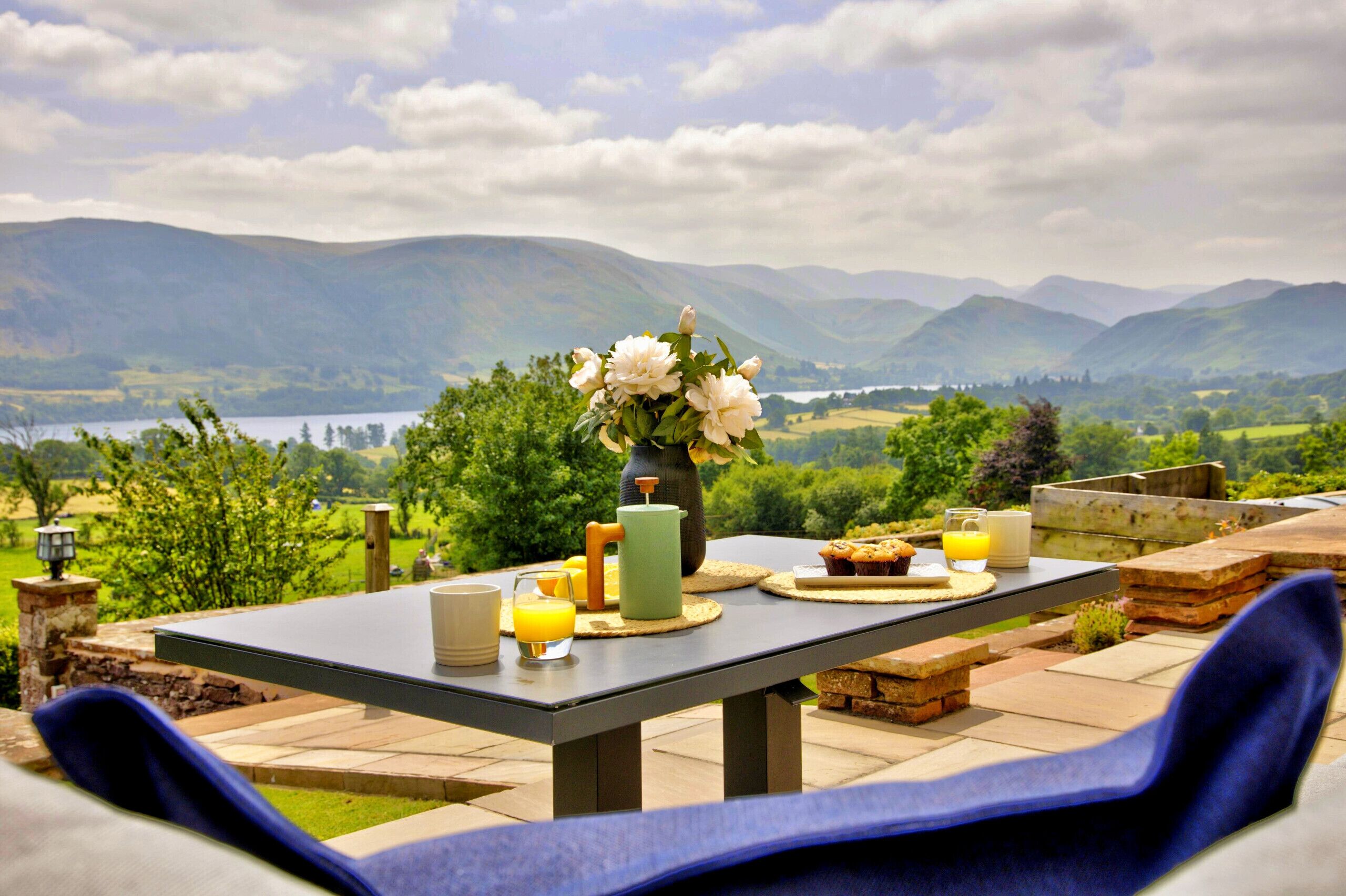 Une table à manger en plein air, avec vue sur les montagnes et un lac, agrémentée d’un vase de fleurs, d’une carafe, d’un verre de jus d’orange et d’une assiette de muffins.