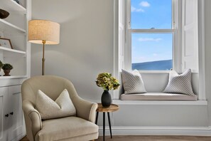 Ein Zimmer mit einem beigen Sessel, einem kleinen Tisch mit Blumenvase, einer Sitzbank mit Kissen und einer Lampe auf einem Regal.