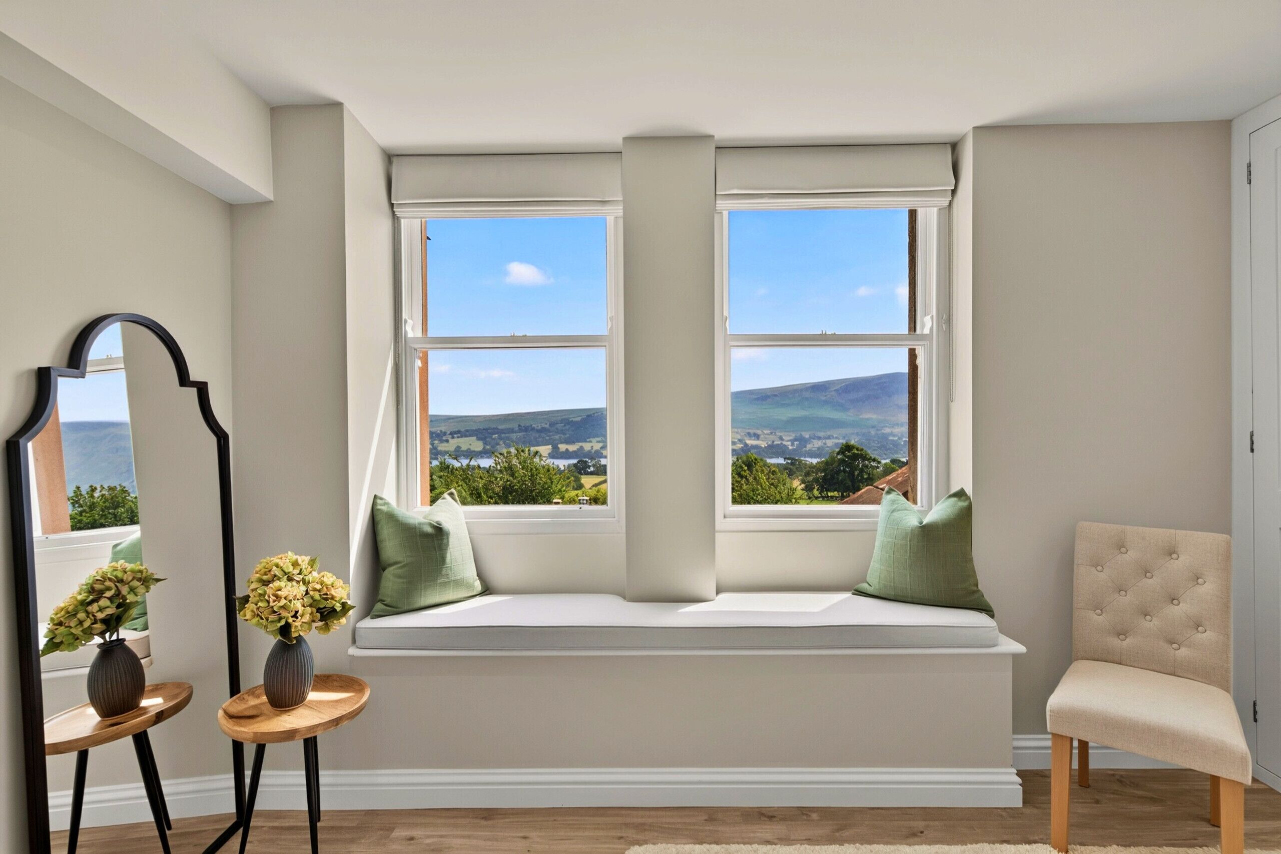 A room with a large window seat, a mirror, a vase with flowers, and a chair.