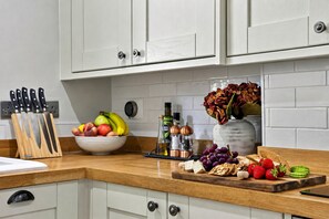 A kitchen with white cabinets, a wooden countertop, a fruit bowl, a cutting board with fruits and cheese, and a vase with flowers. - Willow's View (Wreay)