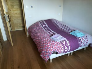 Una cama individual con una colcha estampada, una manta doblada y un almohadón sobre un suelo de madera.