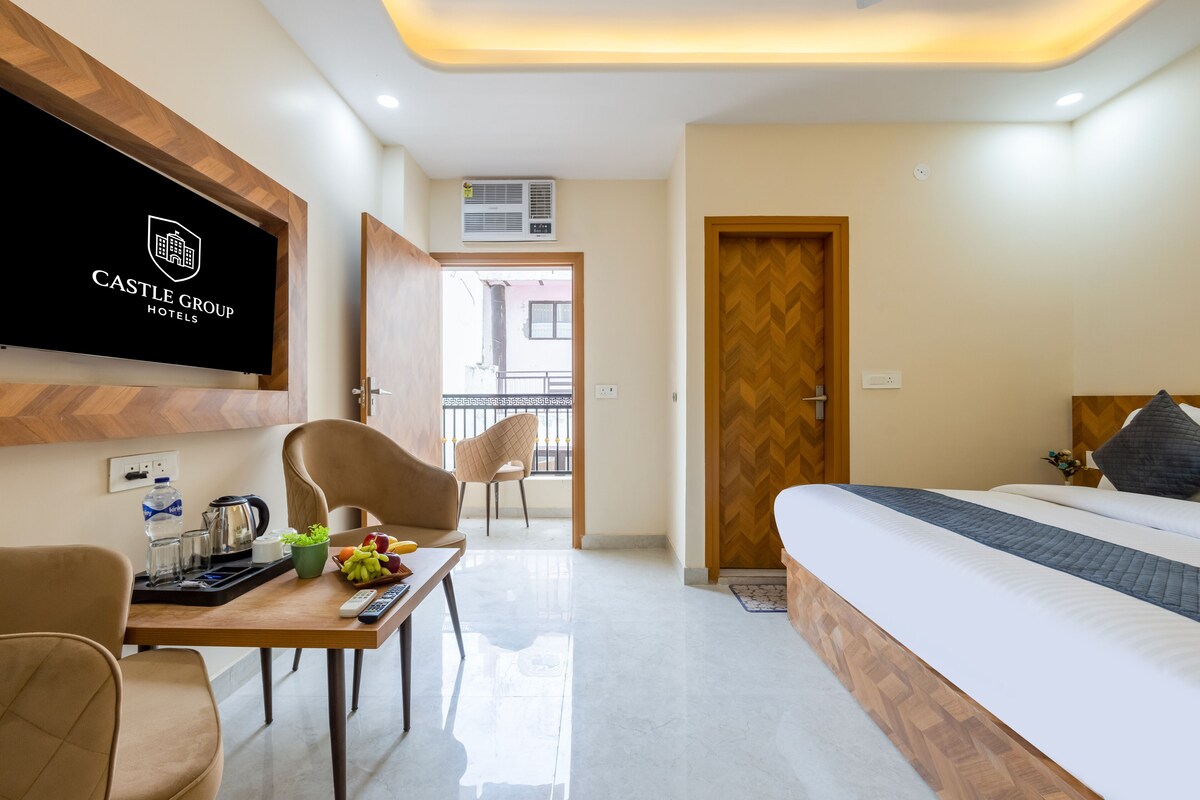 A hotel room with a bed, a TV mounted on the wall, a desk with a kettle and a fruit bowl, and a chair.