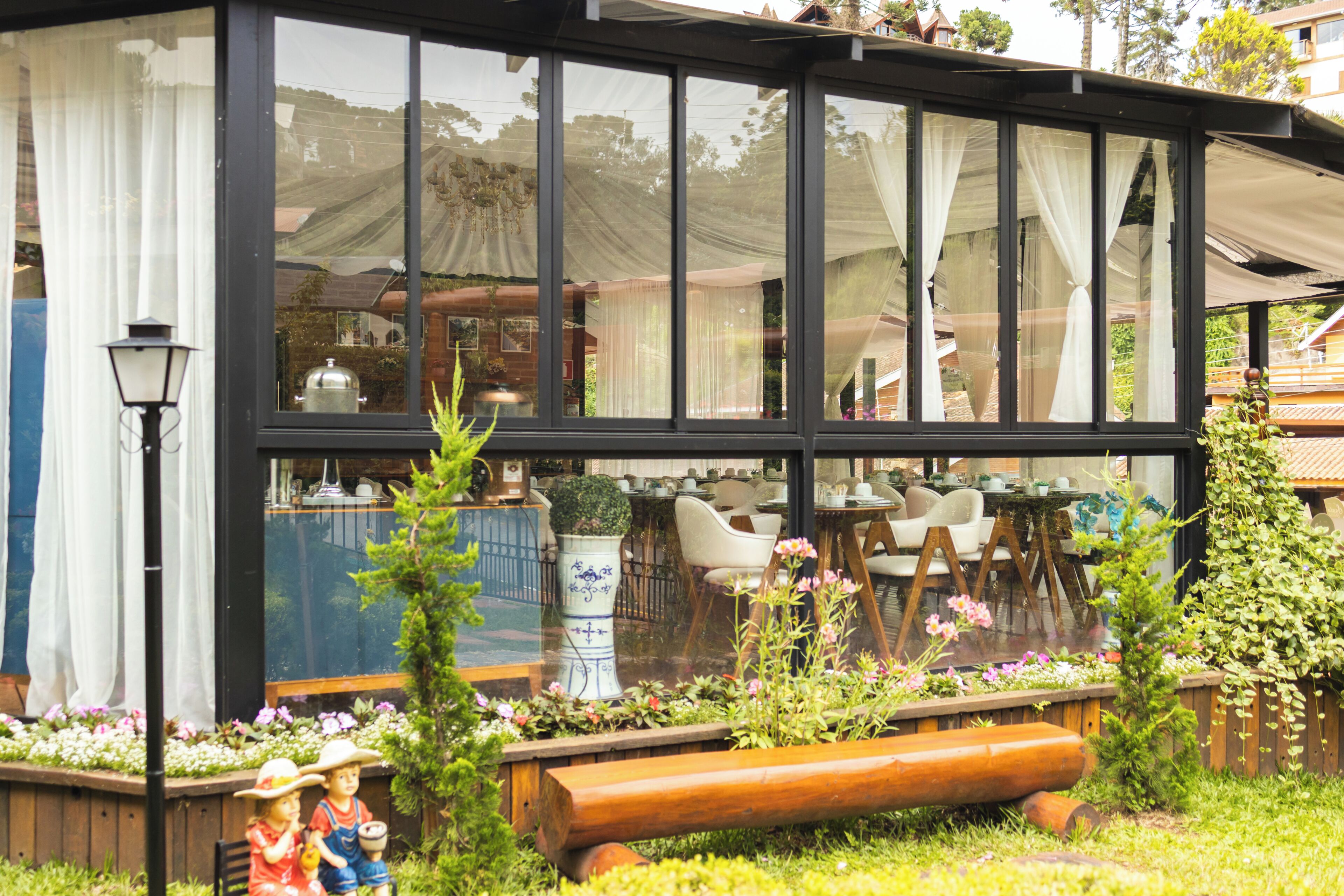 Un restaurante con fachada de vidrio, mesas al aire libre y un jardín con banco de madera y plantas decorativas.