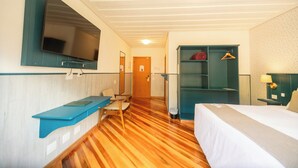 Ett hotellrum med en säng, ett skrivbord med en stol, en tv som är monterad på väggen och en garderob med en spegel.