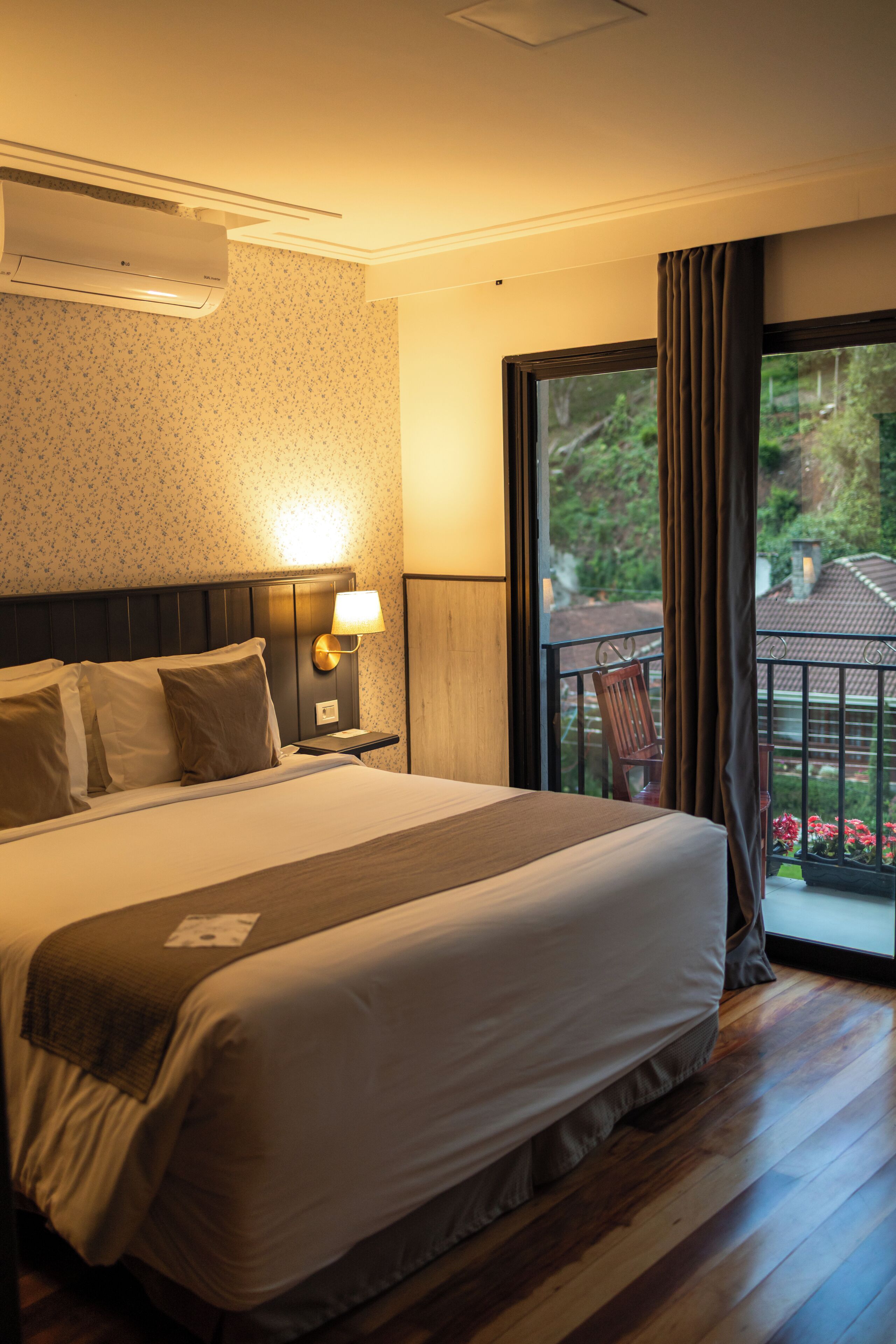 Quarto de hotel com uma cama grande, mesa de cabeceira com abajur, janela com cortinas e vista para o verde lá fora.