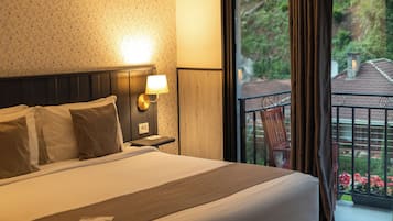 Quarto de hotel com uma cama grande, mesa de cabeceira com abajur, janela com cortinas e vista para o verde lá fora.