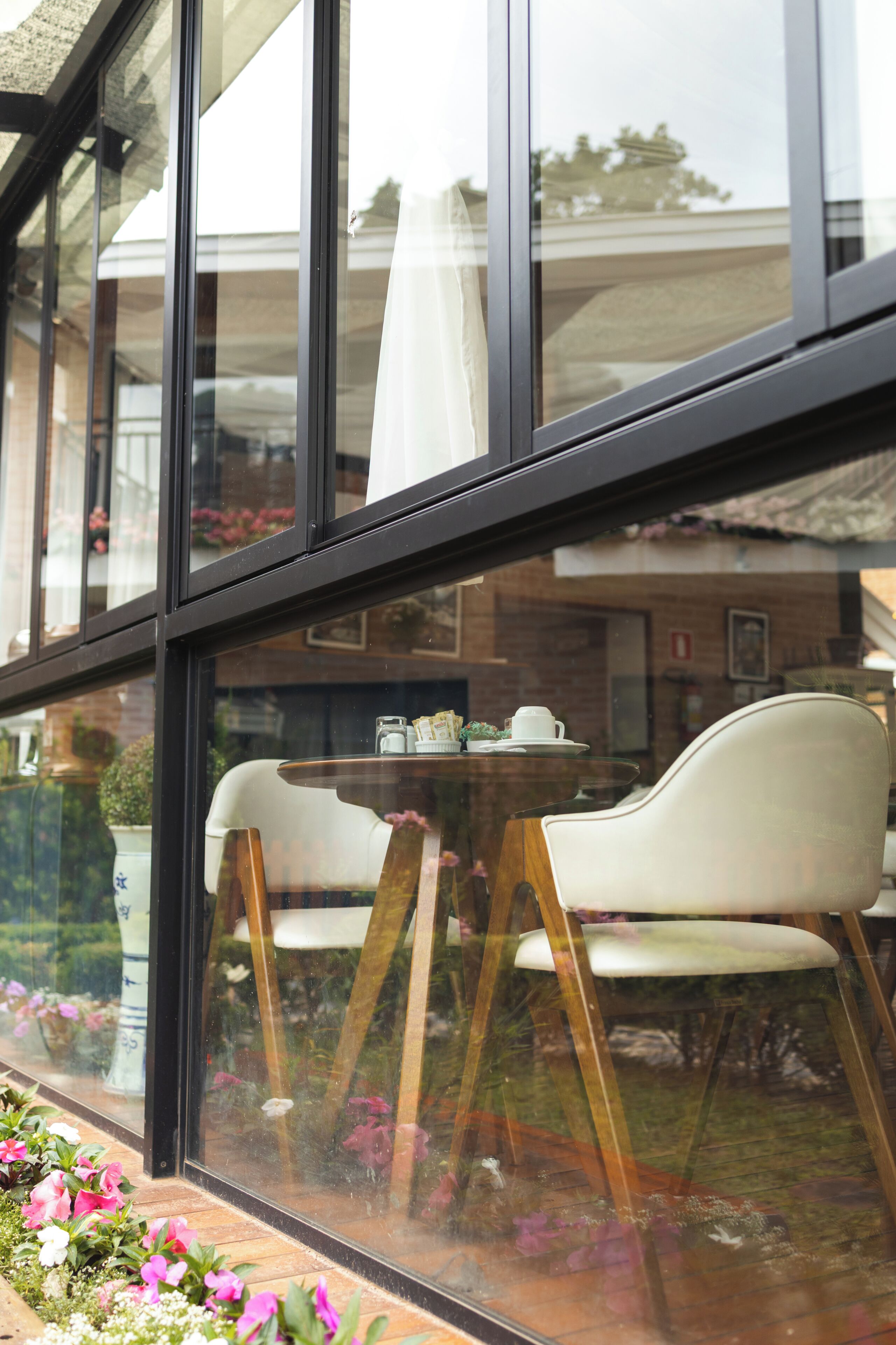 Vista a través de una ventana con una mesa y sillas en el interior, una cortina, y el reflejo de un edificio y vegetación en el exterior.