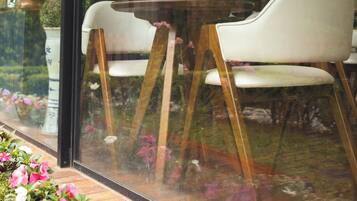 Vista a través de una ventana con una mesa y sillas en el interior, una cortina, y el reflejo de un edificio y vegetación en el exterior.