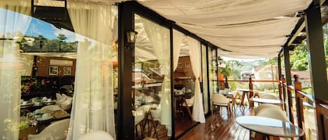 An indoor dining area with wooden tables and chairs, large windows with sheer curtains, and a view of the outdoors.