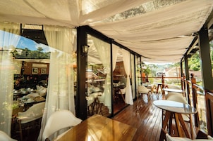 Um espaço de refeições interno com mesas e cadeiras de madeira, grandes janelas com cortinas leves e vista para o exterior.