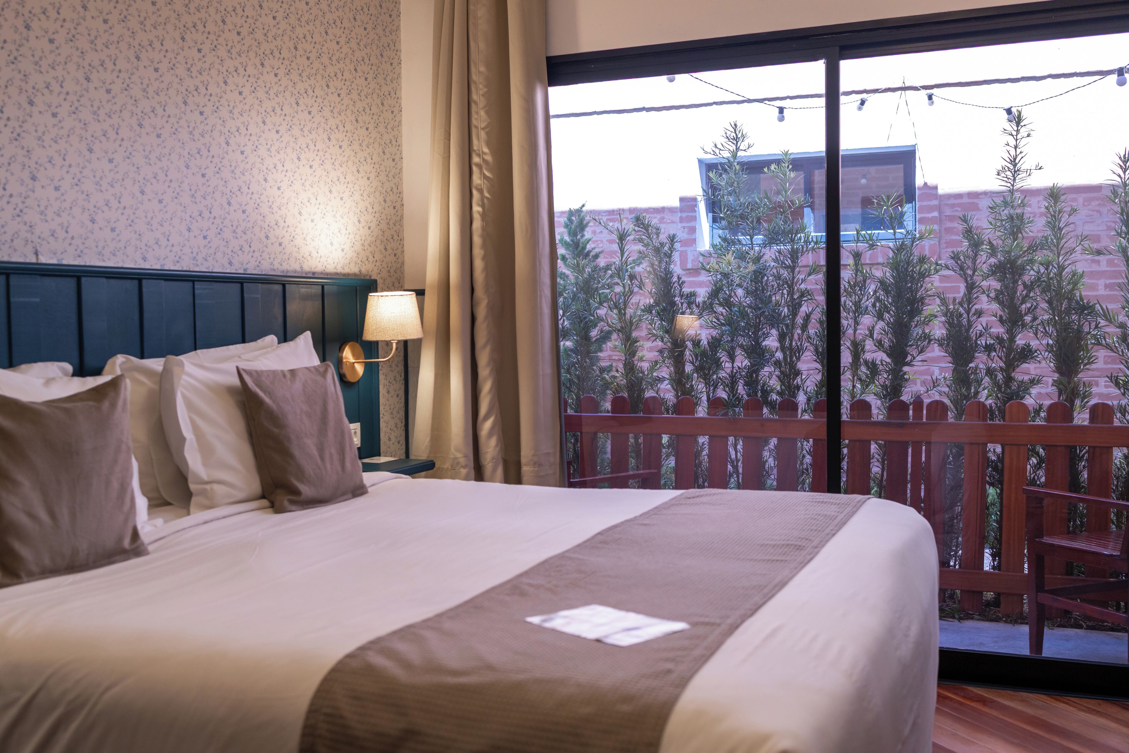 A hotel room with a bed, a nightstand, a lamp, and a view of a balcony with plants and a building.