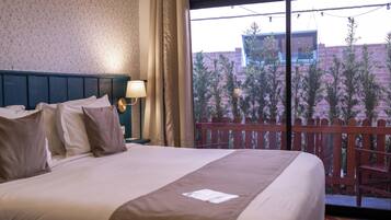 A hotel room with a bed, a nightstand, a lamp, and a view of a balcony with plants and a building.