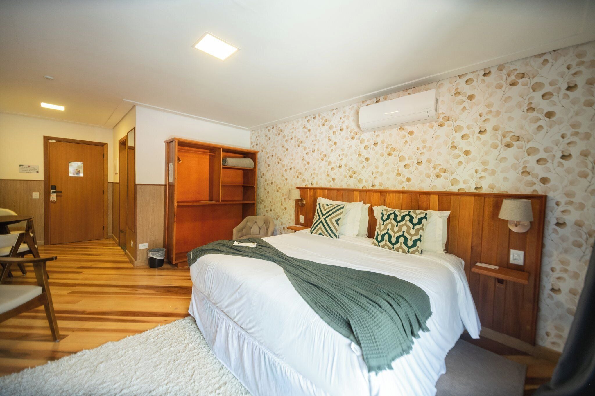 A hotel room with a bed, wooden headboard, a nightstand with a lamp, a wooden wardrobe, and a white carpet.