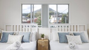 A hotel room with two beds, a wooden nightstand, and a window with a view of houses and hills. - 'Sopra'  Shoal Bay - Close to the beach (Shoal Bay)