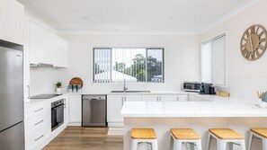 A modern kitchen with white cabinetry, a central island with stools, a stainless steel dishwasher, and a large window with blinds. - 'Sopra'  Shoal Bay - Close to the beach (Shoal Bay)
