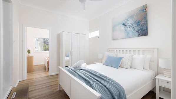 Een slaapkamer met een bed, witte wanden, een plafondventilator, een schilderij en uitzicht op een kamer met een raam.