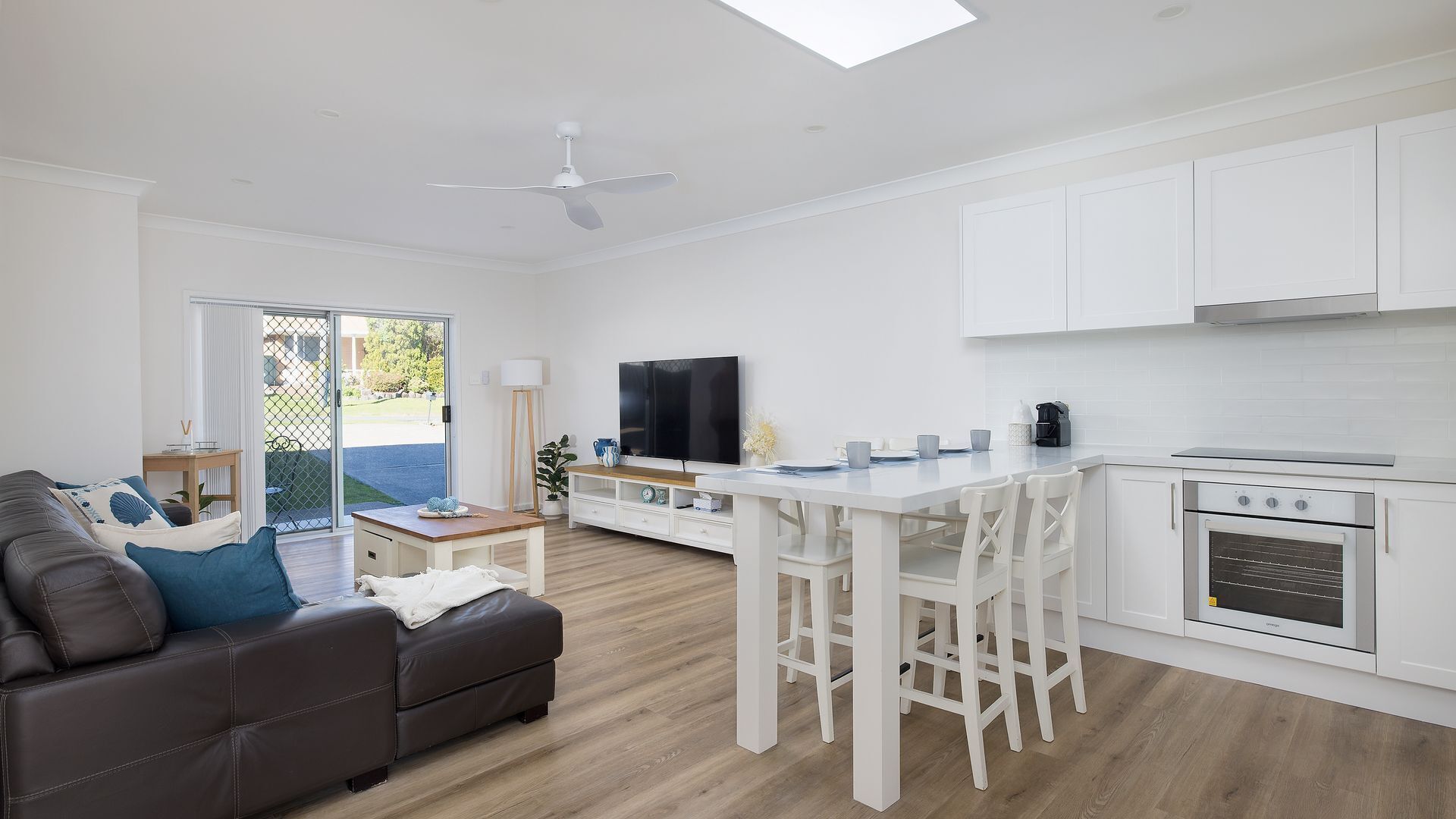 A modern kitchen with white cabinetry, a dining area with white chairs, and a brown leather sofa.