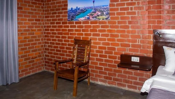 Une pièce avec un mur en briques, une chaise en bois, un lit et un tableau représentant un paysage urbain.