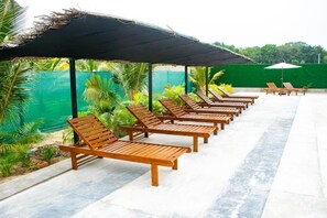 Una fila de sillones de madera bajo una zona sombreada, con una valla verde y palmeras al fondo.