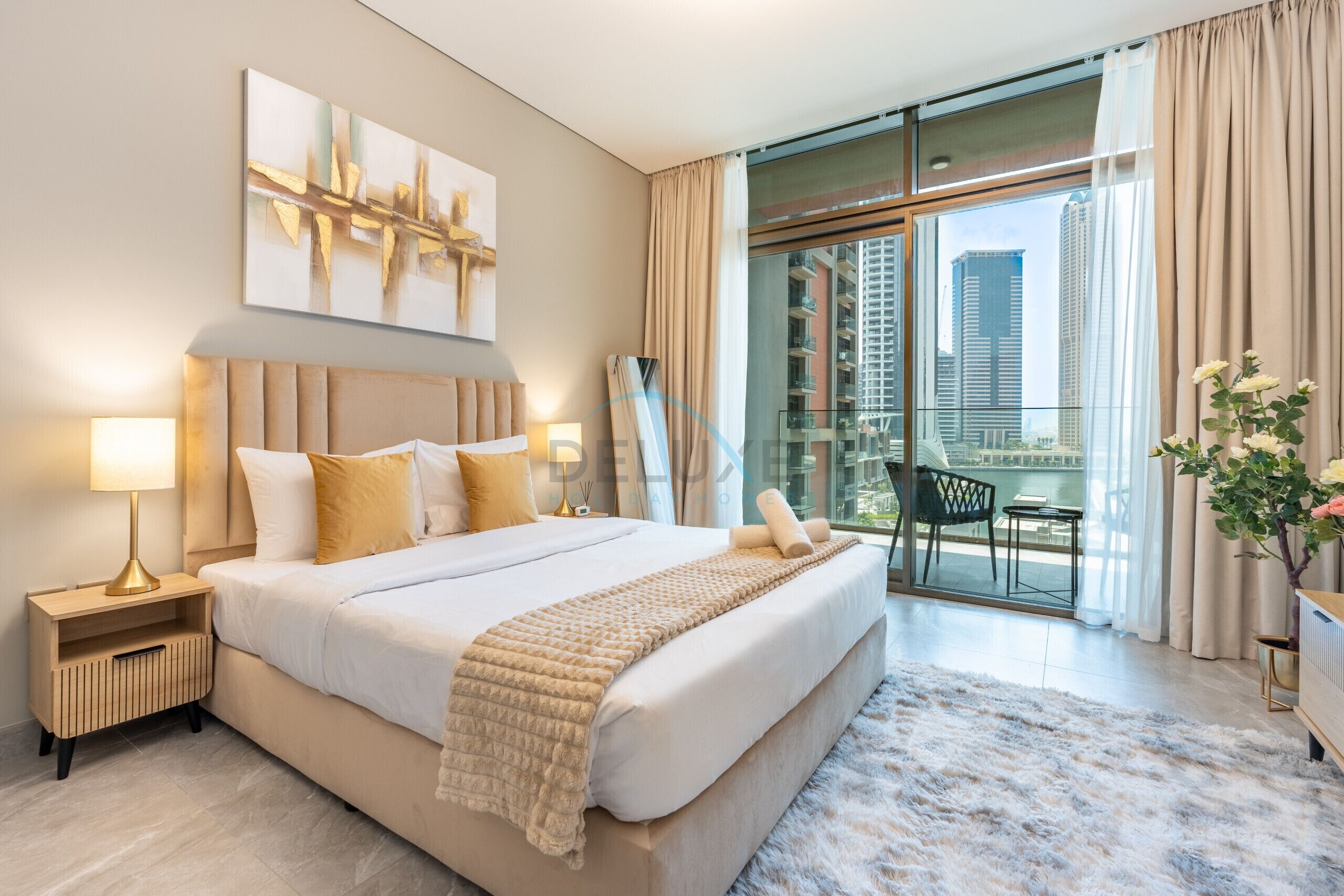 Quarto de hotel moderno com uma cama grande, uma mesa de cabeceira, abajur, vista da cidade por uma varanda e uma planta decorativa.