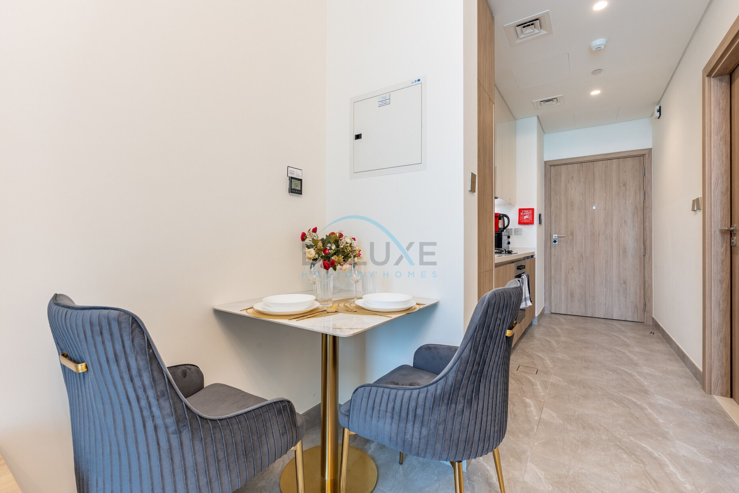 Une chambre d’hôtel avec une table à manger dressée pour deux personnes, deux chaises rembourrées, un petit vase avec des fleurs et un thermostat fixé au mur.