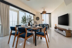 A modern living room with a dining area, large windows, a flat-screen TV, and a chandelier. - High-Floor 2BR with Skyline Views, Moments from Dubai Butterfly Garden (Dubai)