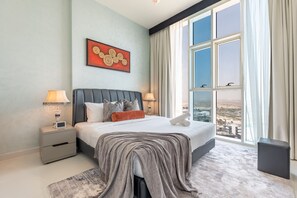 Een moderne slaapkamer met een groot bed, twee nachtkastjes, een bijzettafeltje, uitzicht op de stad en een schilderij aan de muur.