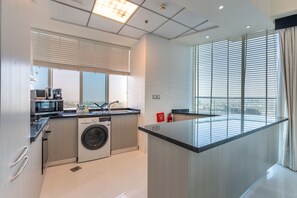 A modern kitchen with a built-in washer and dryer, a large window with blinds, and a view of the cityscape. - High-Floor 2BR with Skyline Views, Moments from Dubai Butterfly Garden (Dubai)