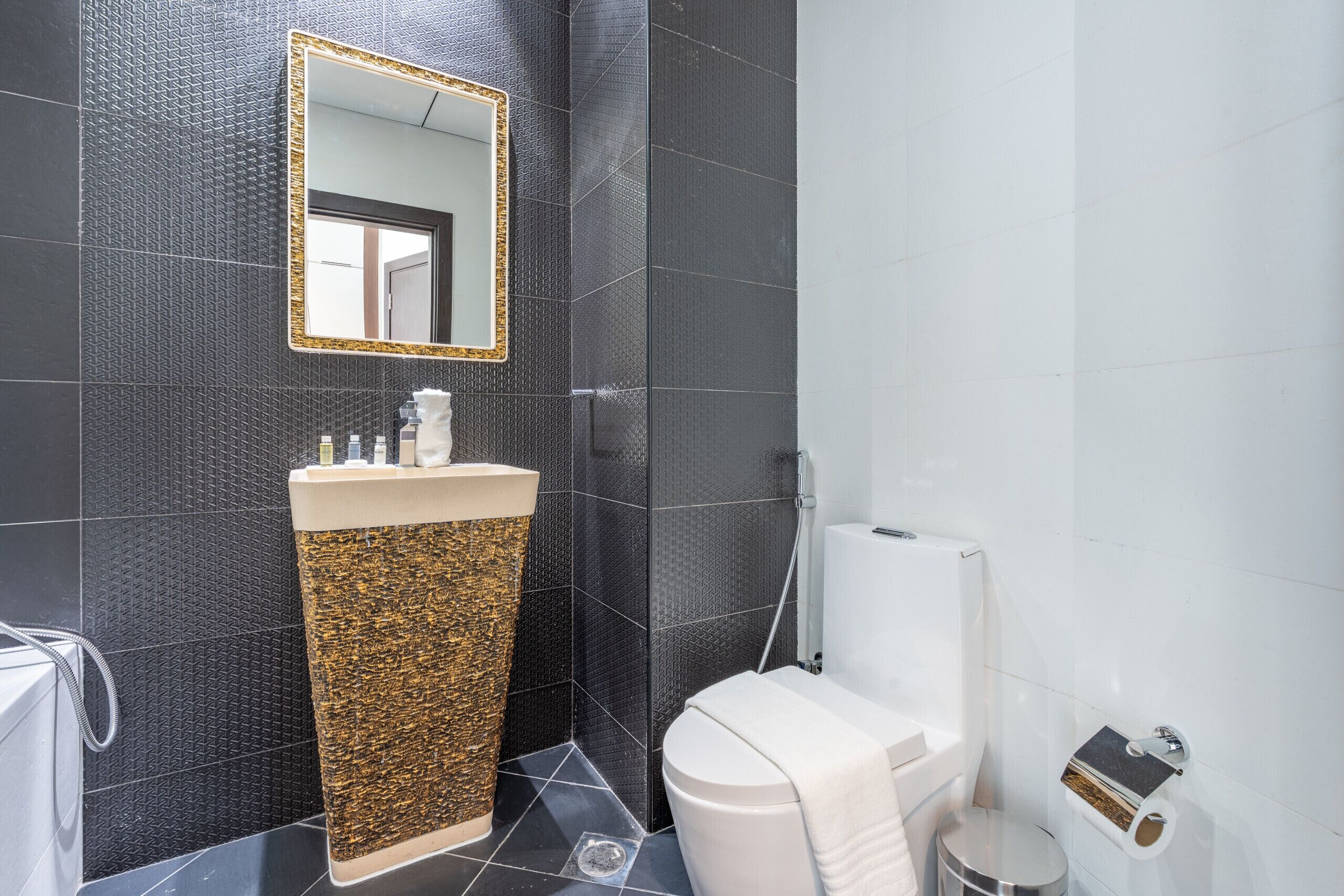 Ein Badezimmer mit schwarzer Wand mit Struktur, einem goldfarbenen und beigefarbenen Waschbecken, einer weißen Toilette und einem Spiegel mit goldener Rahmung.