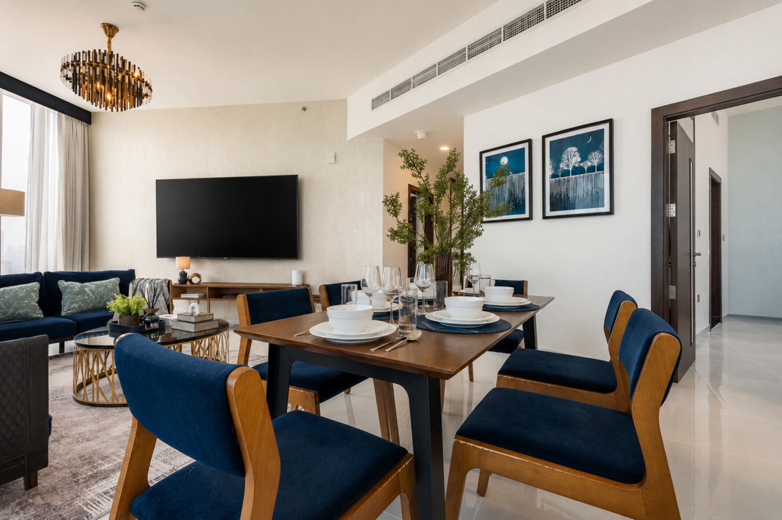 A modern living room with a flat-screen TV, a dining table set for four, and a large window with curtains.