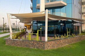 A modern building with a glass facade, a covered outdoor area with exercise equipment, and a brick wall with greenery. - High-Floor 2BR with Skyline Views, Moments from Dubai Butterfly Garden (Dubai)