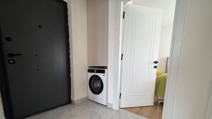 A laundry room with a washing machine, a door, and a hallway leading to a room with a bed. - Qoople Green Ocean A49 - Cozy 1-bedroom apartment in Alanya (Alanya)