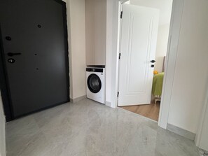 Ein Waschraum mit Waschmaschine, einer Tür und einem Flur, der zu einem Zimmer mit einem Bett führt.
