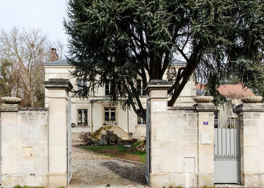 Une entrée fermée par un portail et bordée d’un mur en pierre, un chemin pavé menant à un bâtiment, et un grand arbre au premier plan.