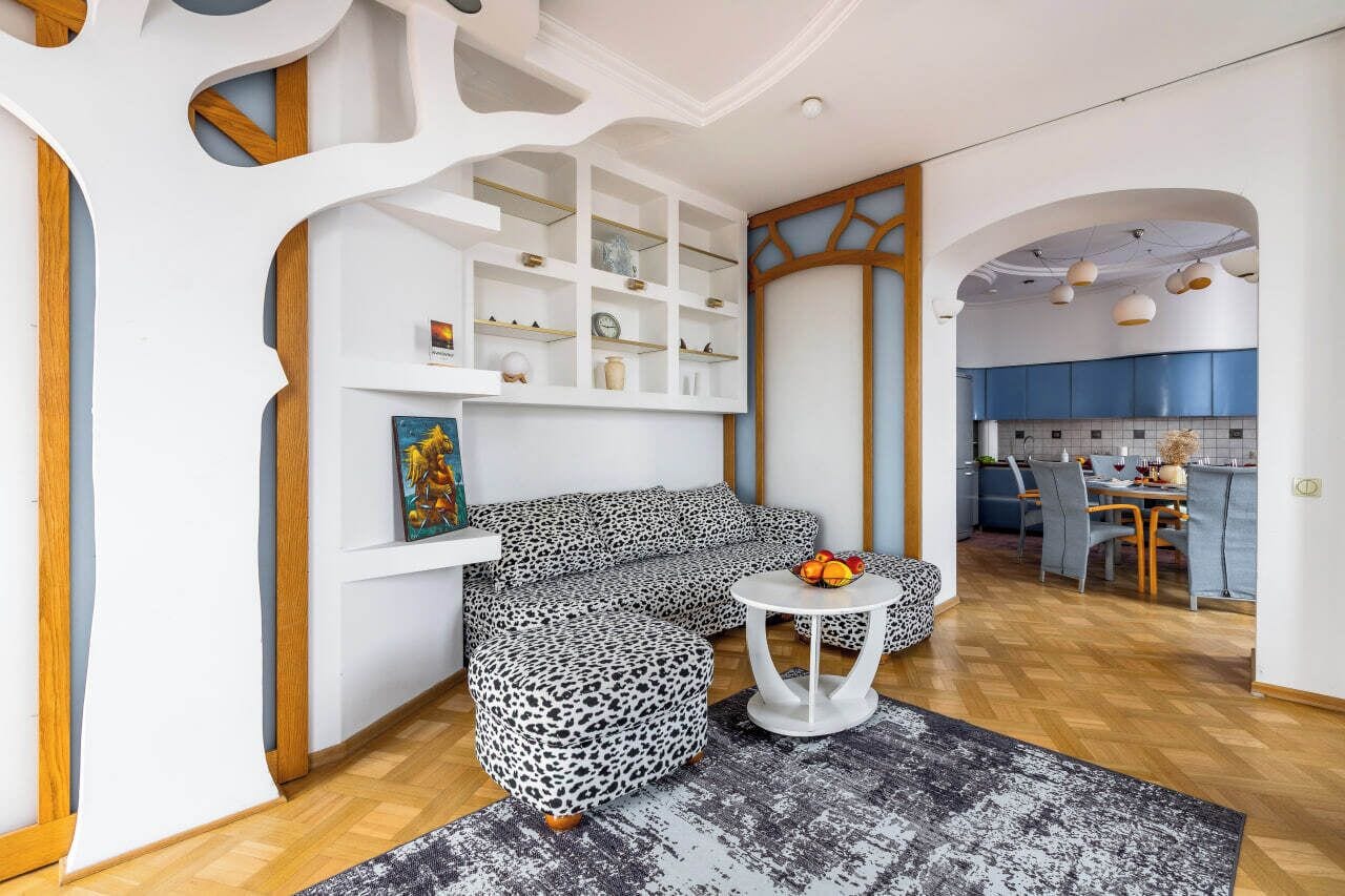 A modern living room with a sofa, ottoman, and a small table with fruit. There is a kitchen visible in the background.
