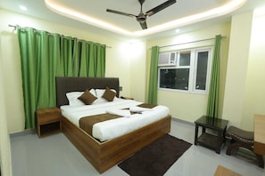 A bedroom with a bed, a nightstand, a bedside table, a ceiling fan, a window with curtains, and an air conditioning unit. - Hill View Haridwar Run by R.K. Group (Haridwar)