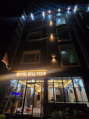 Een hotelentree met een bord waarop 'HOTEL HILL VIEW' staat en een receptiebalie zichtbaar achter de glazen deuren.