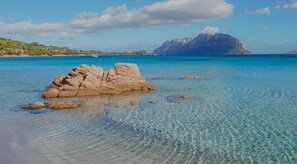 Klares blaues Meer mit einem Felsen im Vordergrund und einem Berg im Hintergrund.