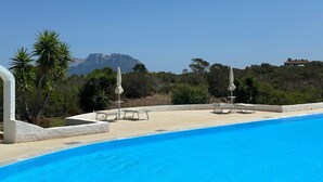 Una piscina con sillas de descanso y sombrillas, rodeada de follaje y una montaña al fondo.