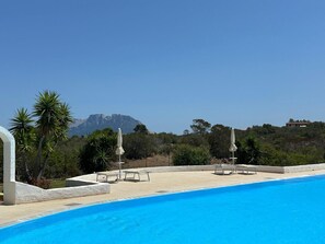Ein Schwimmbad mit Liegestühlen und Sonnenschirmen, umgeben von Grünflächen und einem Berg im Hintergrund.